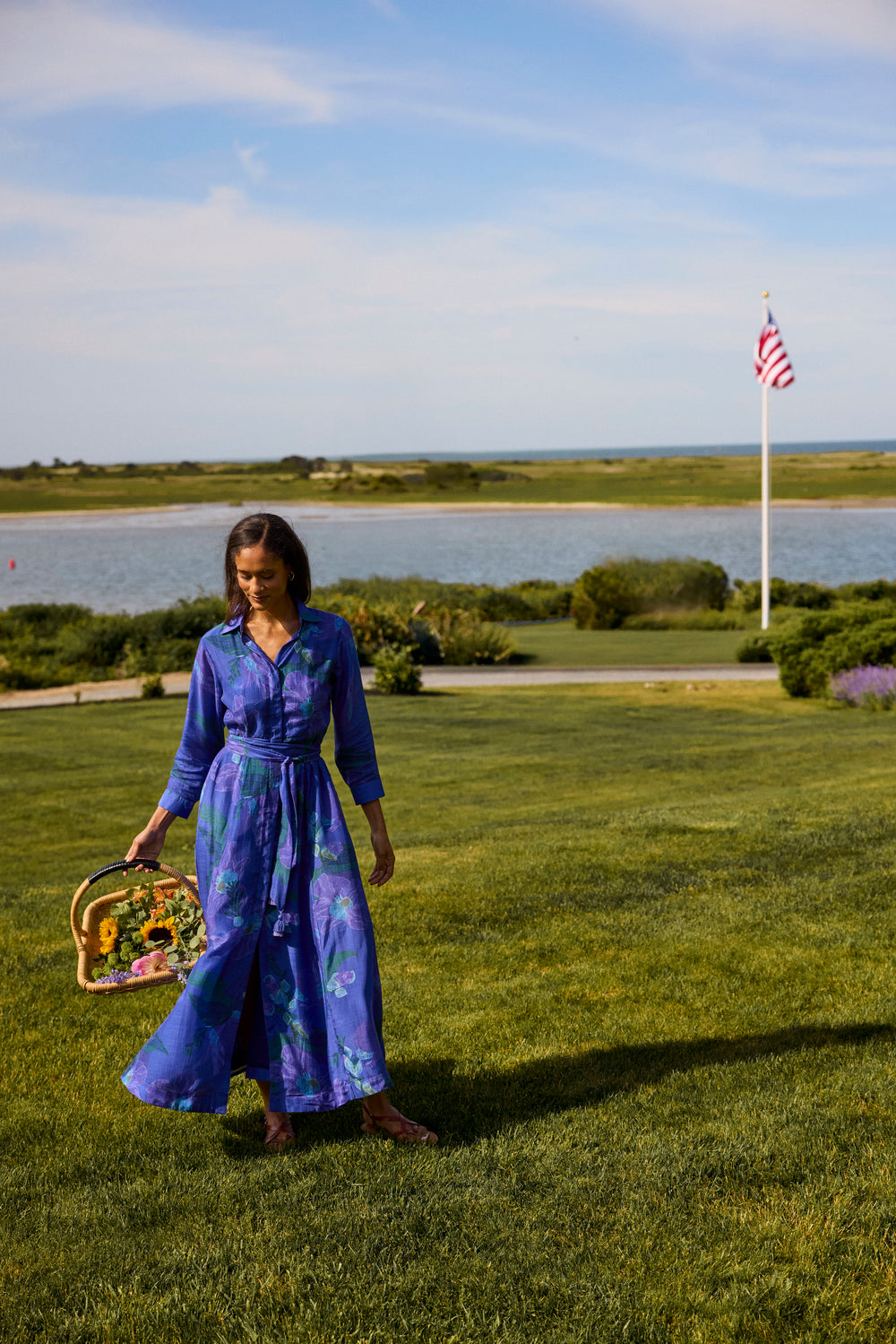 Kit Shirtdress in Helin Royal, front view on a lawn by the water, carrying a flower basket